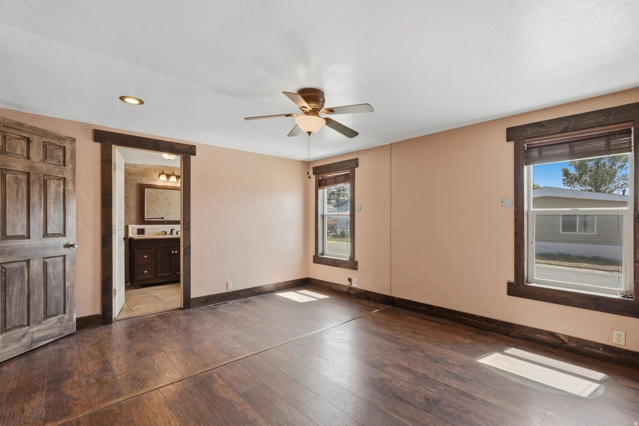 Spacious room featuring wood-finish flooring, two large windows with dark wood trim, a ceiling fan with integrated lighting, and a recessed ceiling light