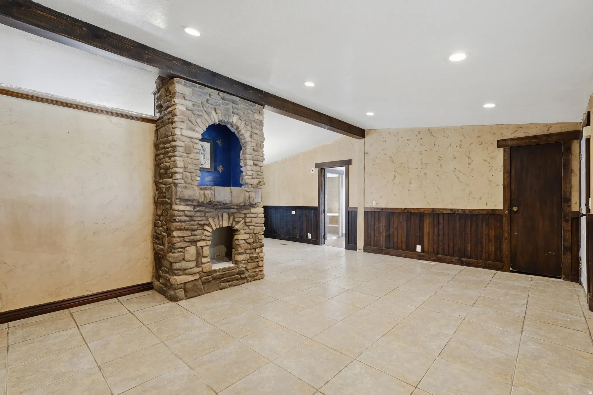 Stone-clad fireplace with an arched niche, tile flooring, wood paneling, and exposed ceiling beams