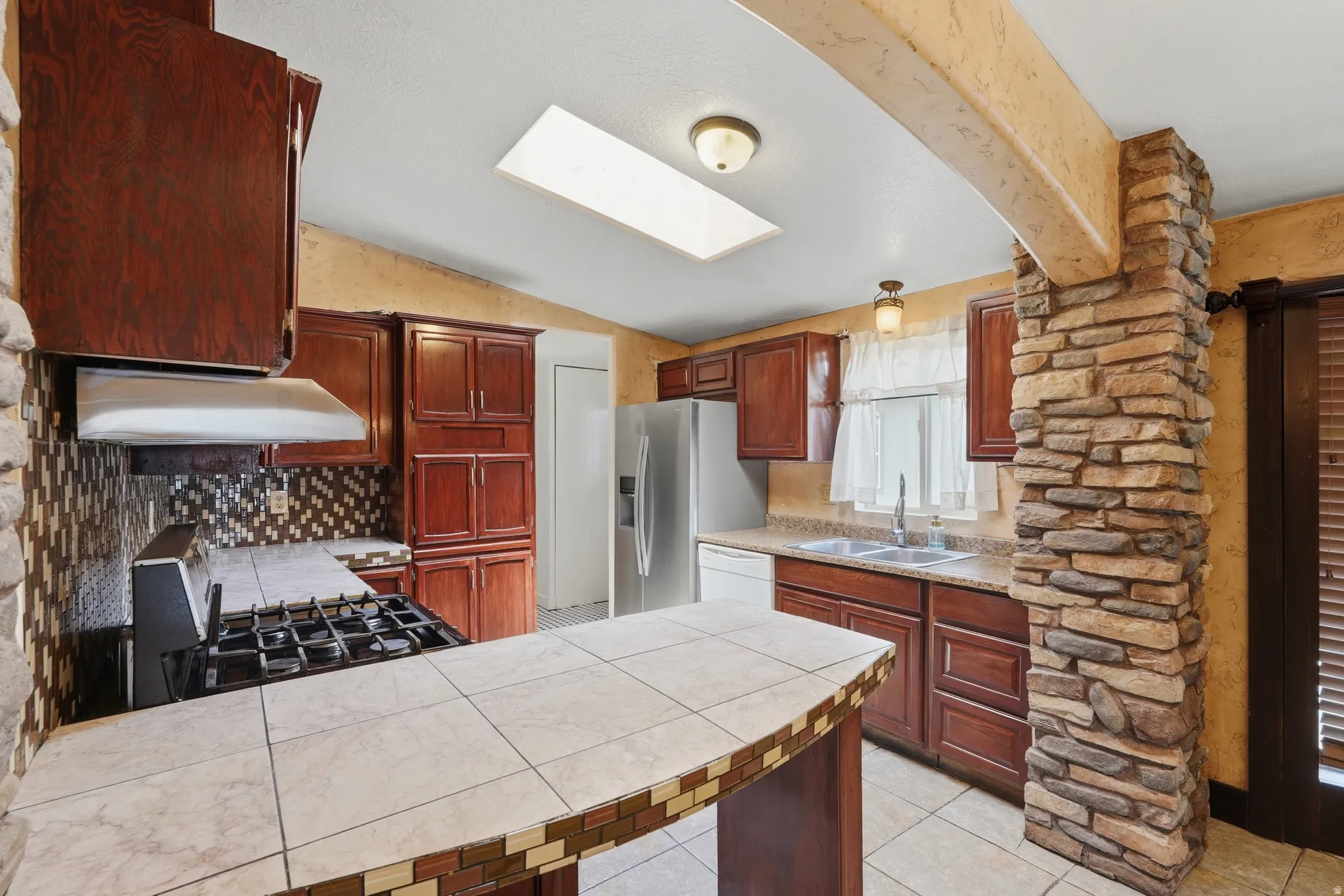 Kitchen featuring a stone-clad pillar, wood cabinetry, tile countertops, and a gas range with an overhead vent