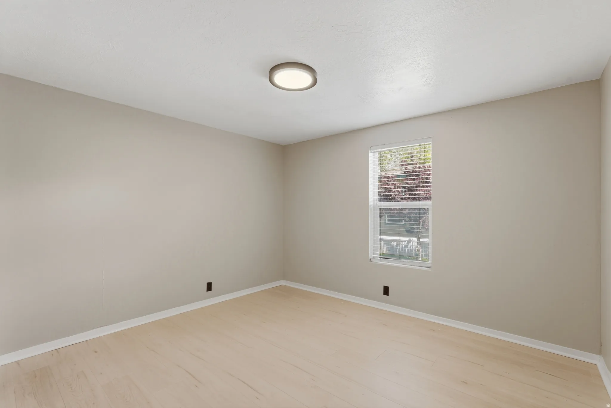 Neutral-toned room featuring a single window with blinds, light wood-finish flooring, and a flush-mount ceiling light fixture
