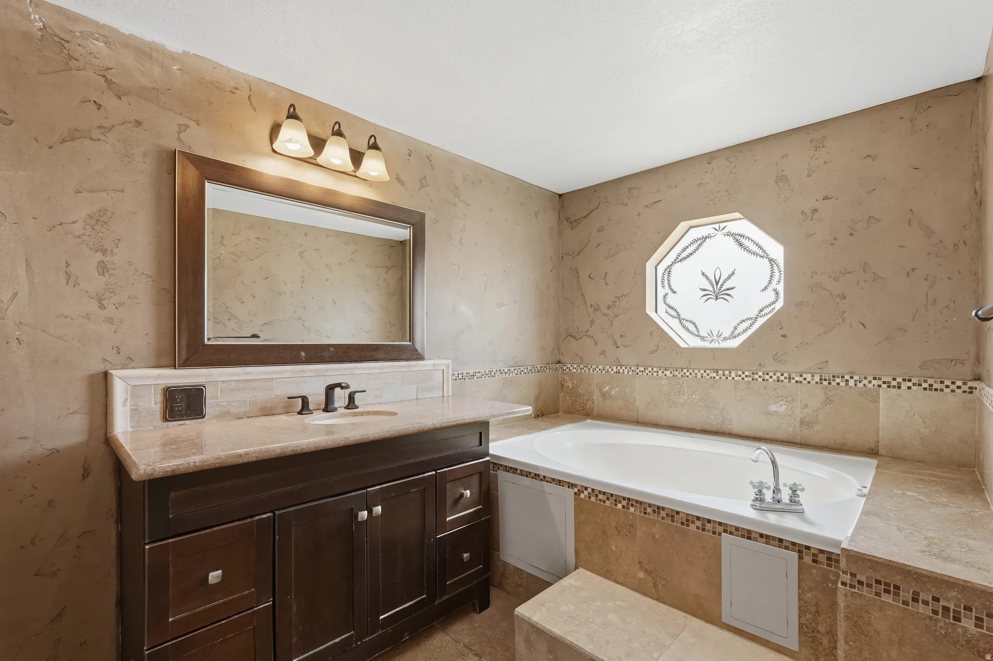 Built-in soaking tub with mosaic tile accents, octagonal frosted window, dark wood-finish vanity with stone countertop, framed mirror, and a three-light fixture