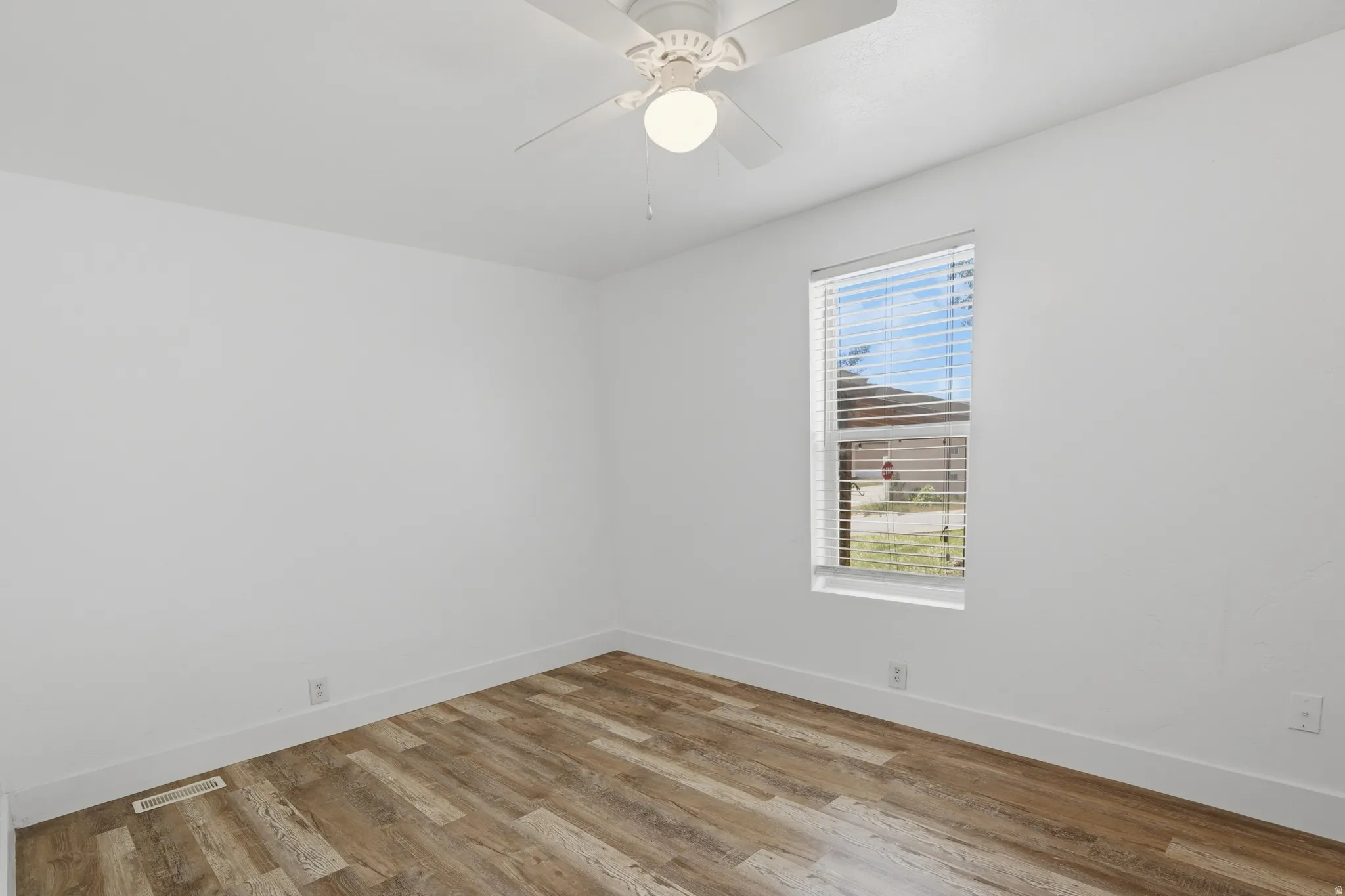 Bright interior space featuring wood-finish flooring, a single window with blinds, and a white ceiling fan with integrated lighting