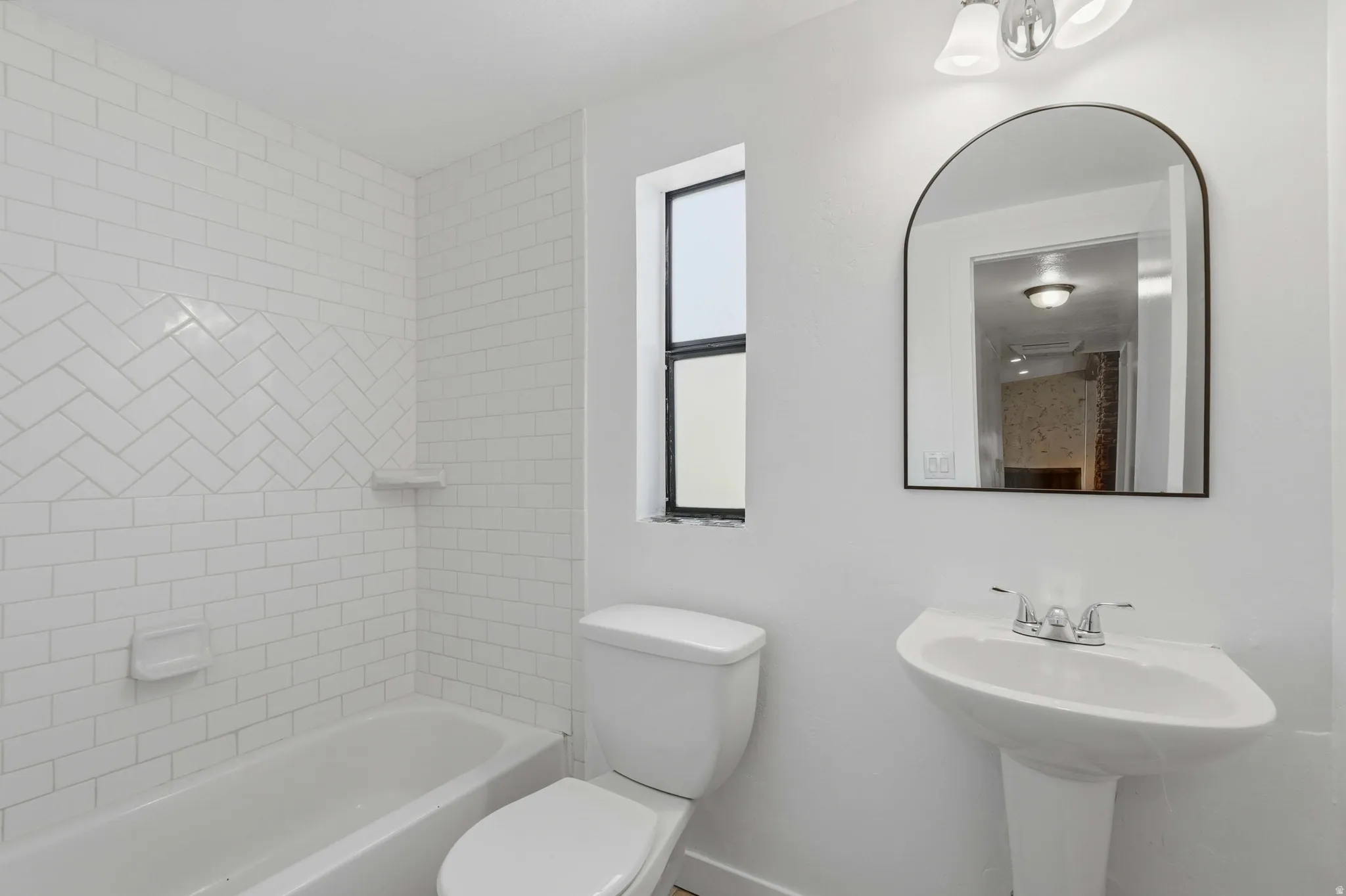 White-tiled bathroom featuring a pedestal sink with a chrome faucet, an arched mirror, and a bathtub with subway tile surround and a herringbone accent band
