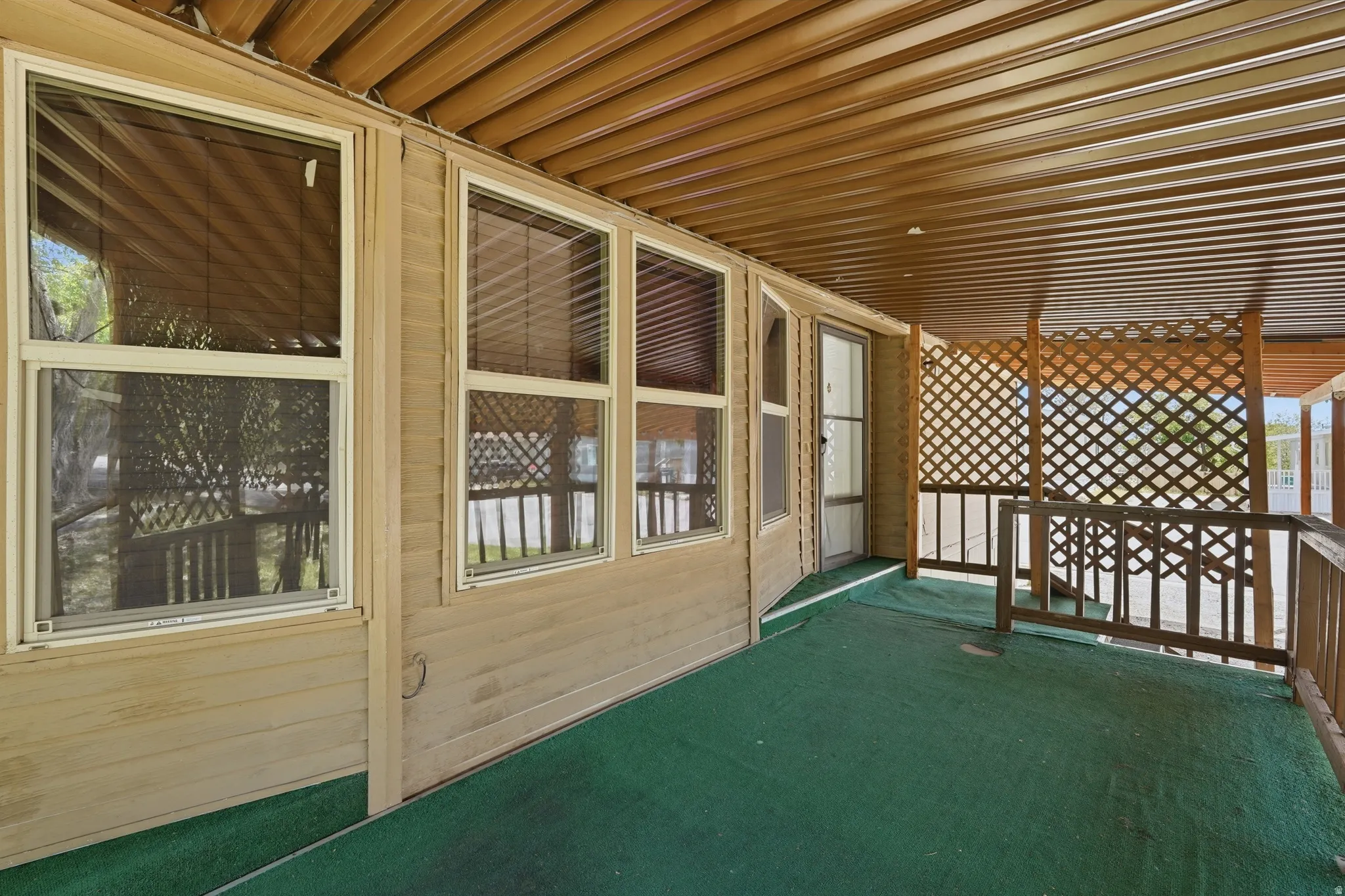 Covered patio area featuring a green artificial turf floor, lattice privacy screening, multiple windows with blinds, and a paneled ceiling