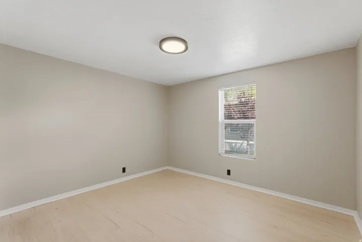 Neutral-toned room featuring a single window with blinds, light wood-finish flooring, and a flush-mount ceiling light fixture