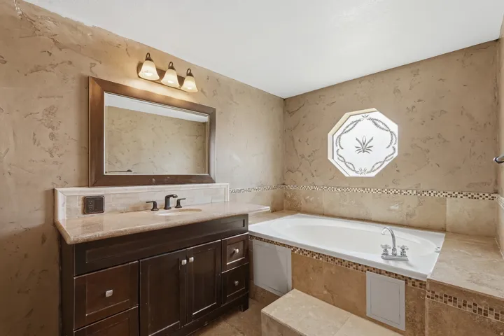 Built-in soaking tub with mosaic tile accents, octagonal frosted window, dark wood-finish vanity with stone countertop, framed mirror, and a three-light fixture