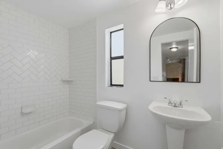 White-tiled bathroom featuring a pedestal sink with a chrome faucet, an arched mirror, and a bathtub with subway tile surround and a herringbone accent band