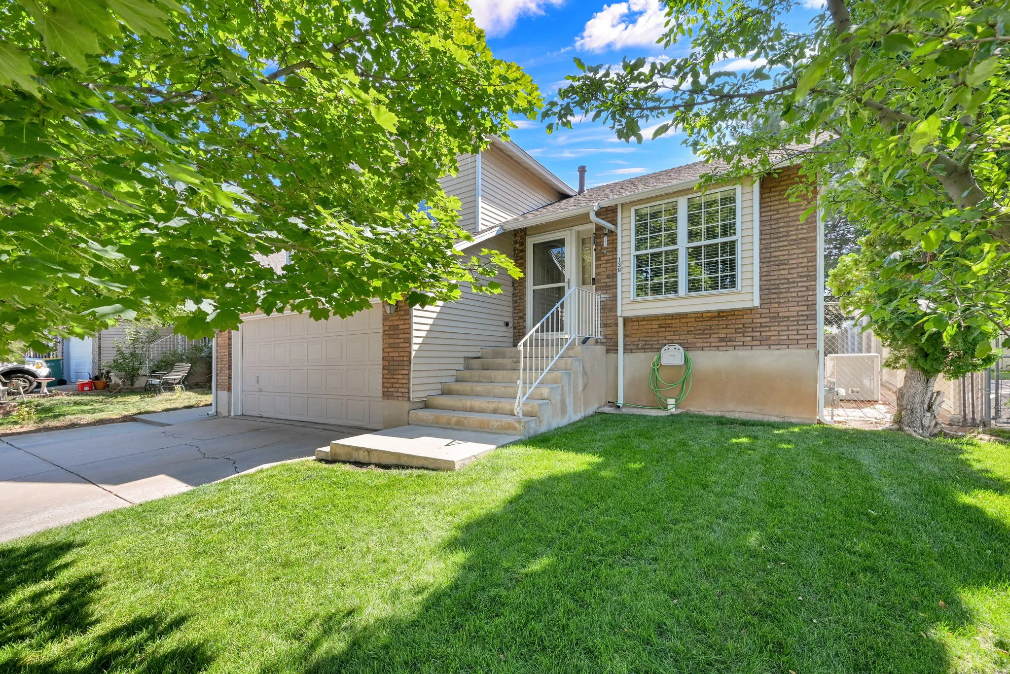 Brick-front home featuring a manicured lawn, mature shade trees, attached two-car garage, and a welcoming front entrance with white railings and stone steps