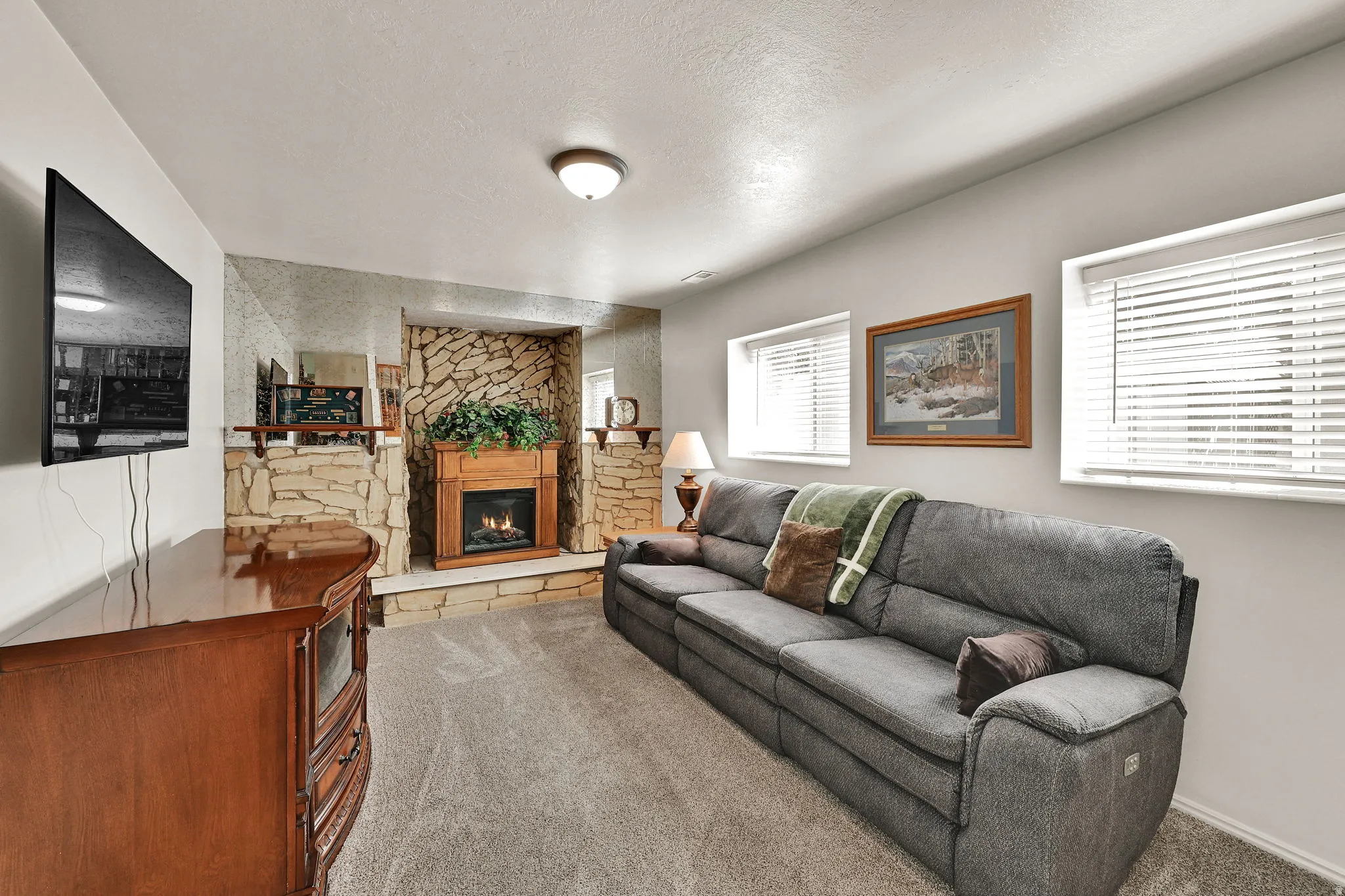 Living area featuring a stone-clad fireplace surround with a wood mantel, soft gray carpeting, and two windows with horizontal blinds