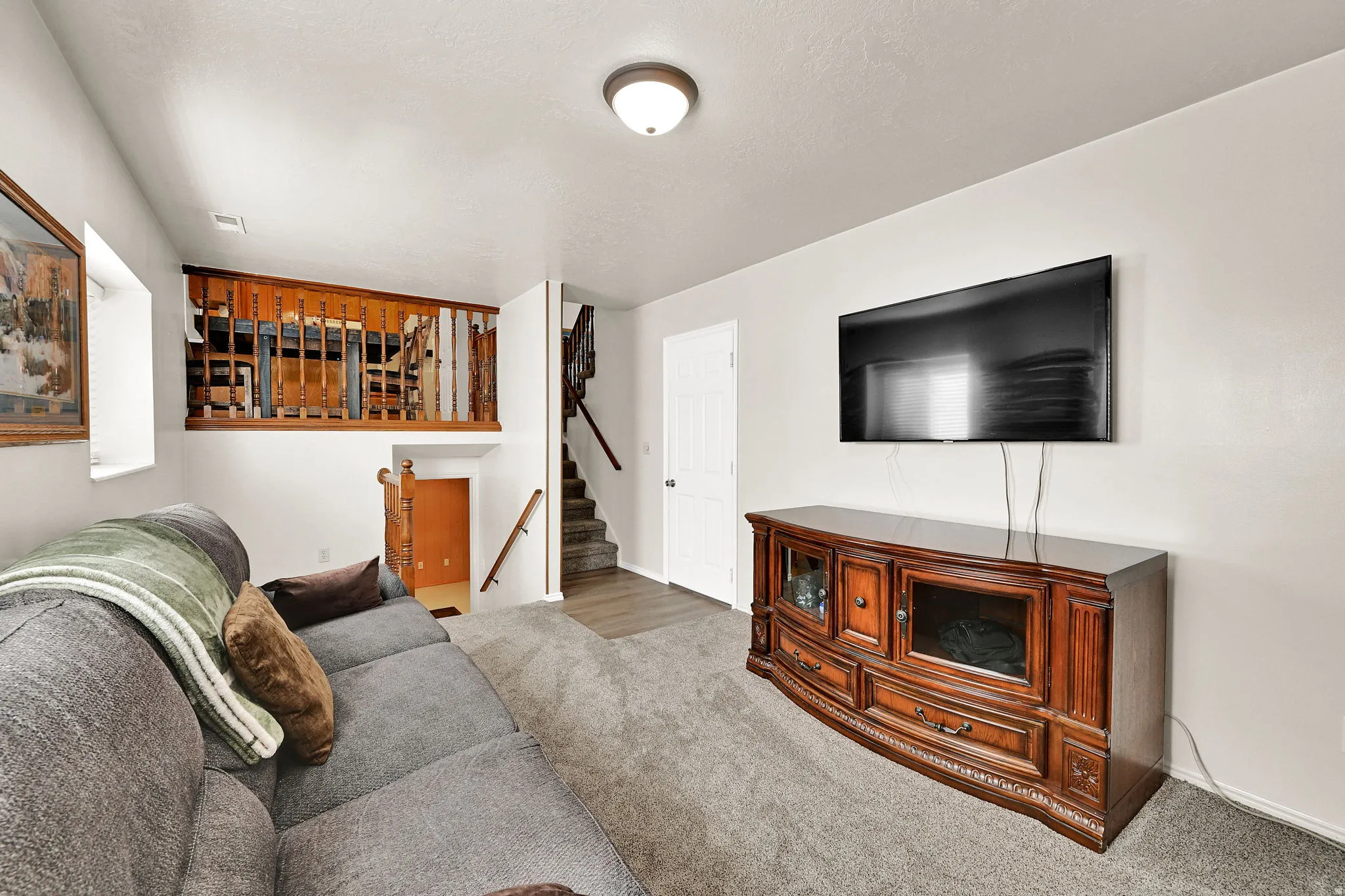 Finished basement recreation room featuring a gray carpeted floor, wall-mounted flat-screen television above a wood-finish media console with storage cabinetry, gray upholstered seating, and recessed ceiling lighting