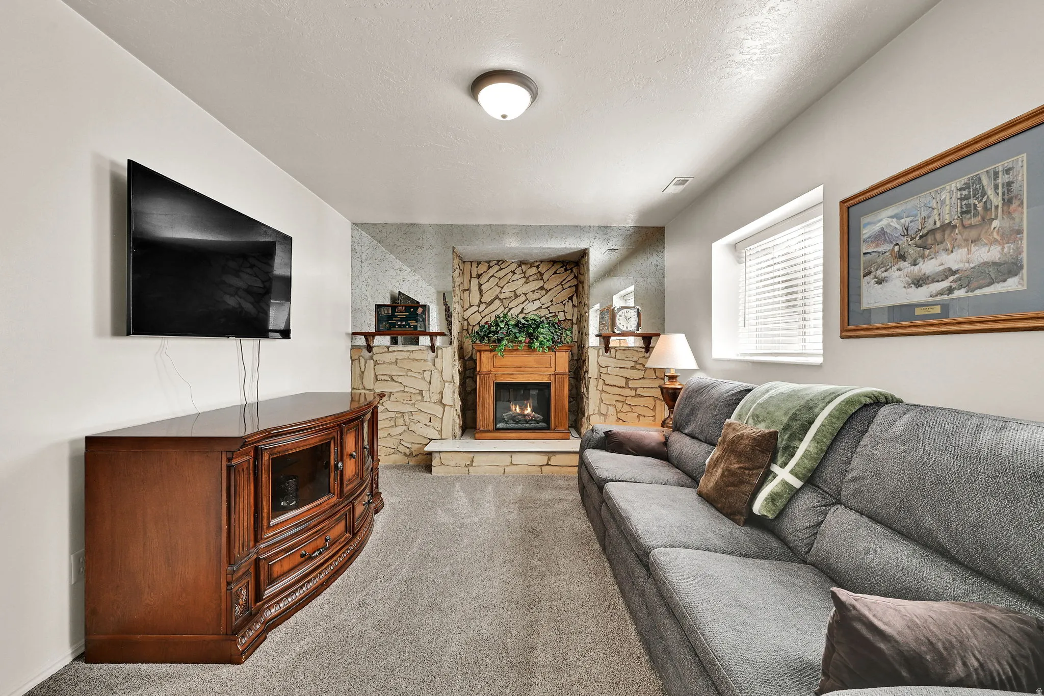 Finished basement recreation room featuring a wood-finish media console with built-in cabinetry, wall-mounted flat-screen television, stone fireplace with wood mantel, gray upholstered sectional sofa, recessed ceiling lighting, and carpet flooring through