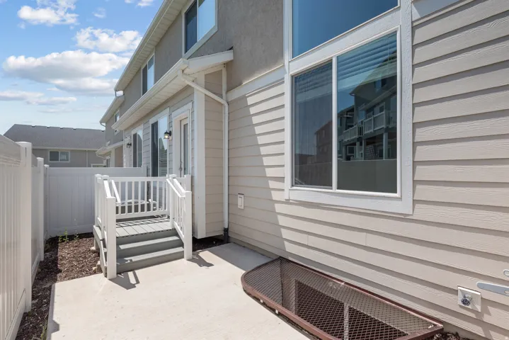 Rear exterior featuring light-colored siding and stucco, a private patio, white vinyl fencing, a multi-pane window, and a ground-level basement window well with a metal grate