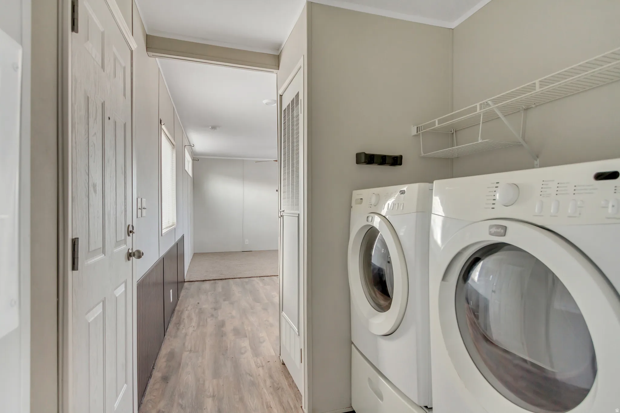 Dedicated laundry room featuring side-by-side front-load washer and dryer with white finish, overhead shelving for storage, wood-finish flooring, and neutral wall tones