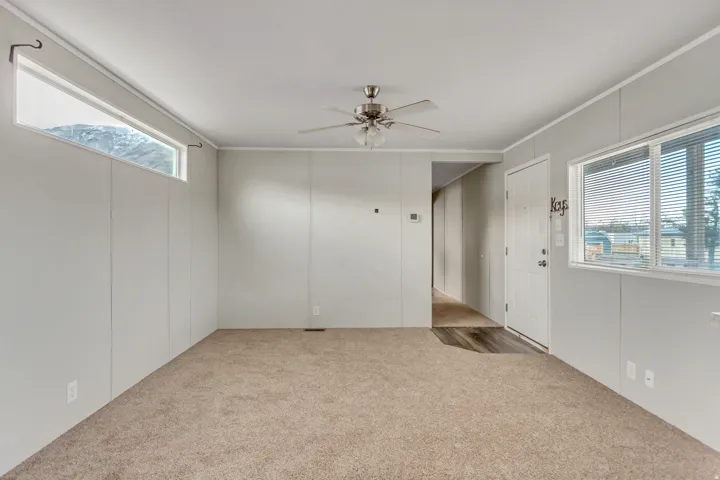 Carpeted living area featuring a ceiling fan with integrated lighting, a front entry door with an adjacent window, and a high-set transom window