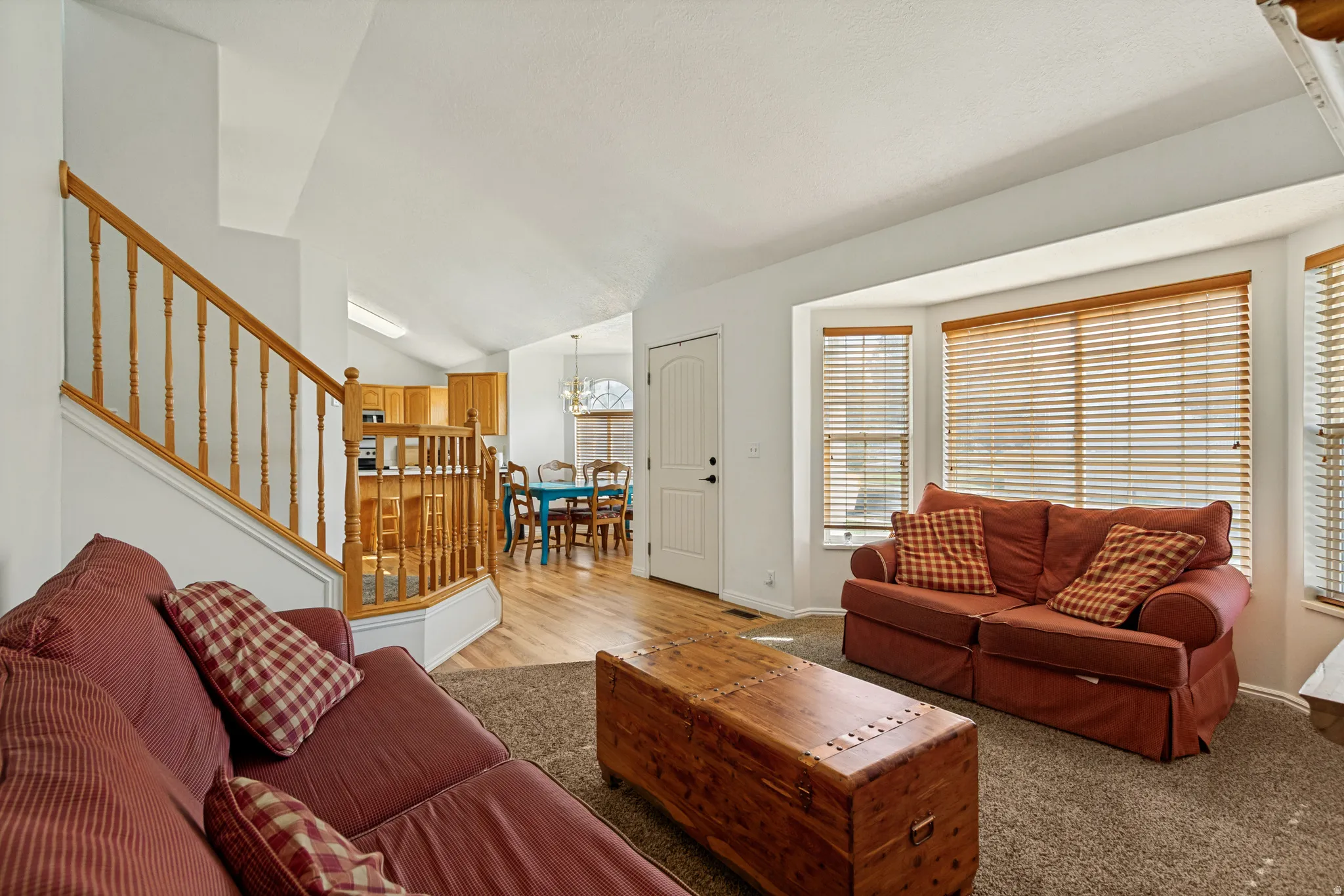 Living area featuring a bay window with natural wood blinds, vaulted ceiling, and carpeted flooring