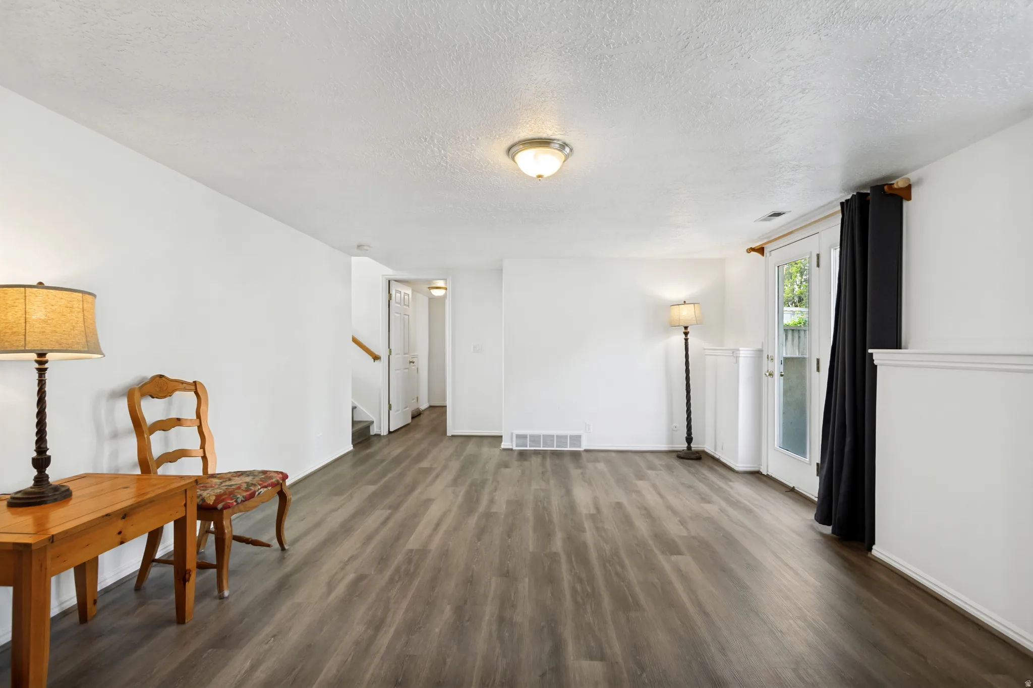 Spacious open-concept living area showcasing wood-finish flooring throughout, abundant natural light from multiple windows and glass doors, neutral white walls, and ceiling-mounted lighting fixtures