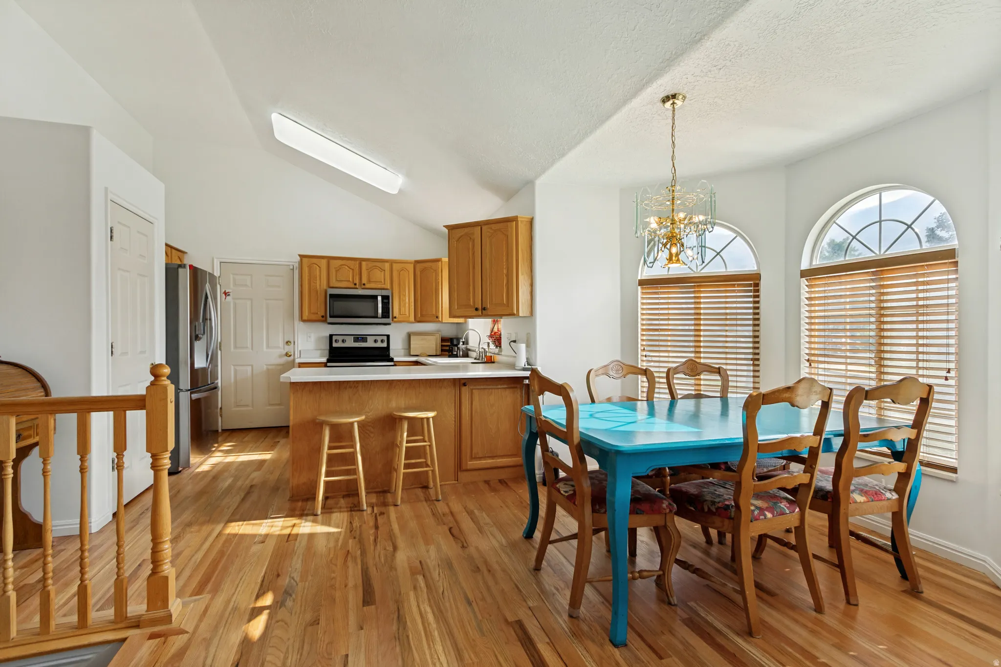 Open-concept kitchen and dining area featuring warm wood-finish flooring throughout, custom cabinetry with stainless steel appliances, a centered island with seating, and abundant natural light from arched windows with plantation shutters