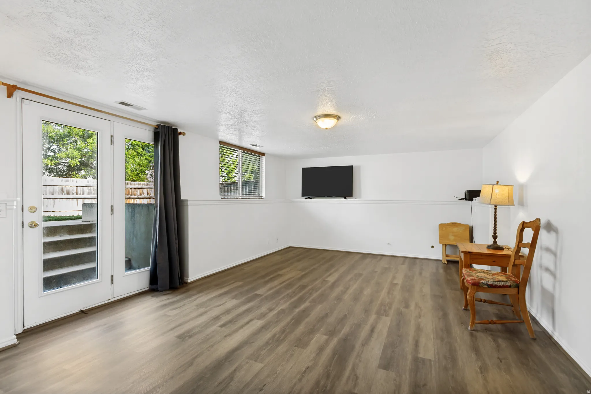 Spacious recreation room featuring wood-finish flooring throughout, abundant natural light from multiple windows and glass door access to exterior space, and white walls with wainscoting detail