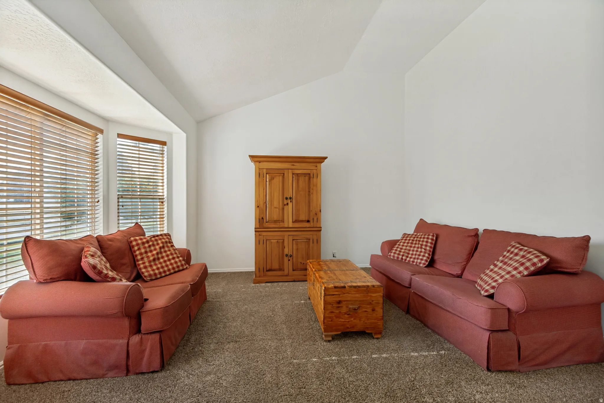 Spacious living area featuring vaulted ceilings, large multi-pane windows with plantation shutters, neutral carpet flooring, and substantial seating capacity
