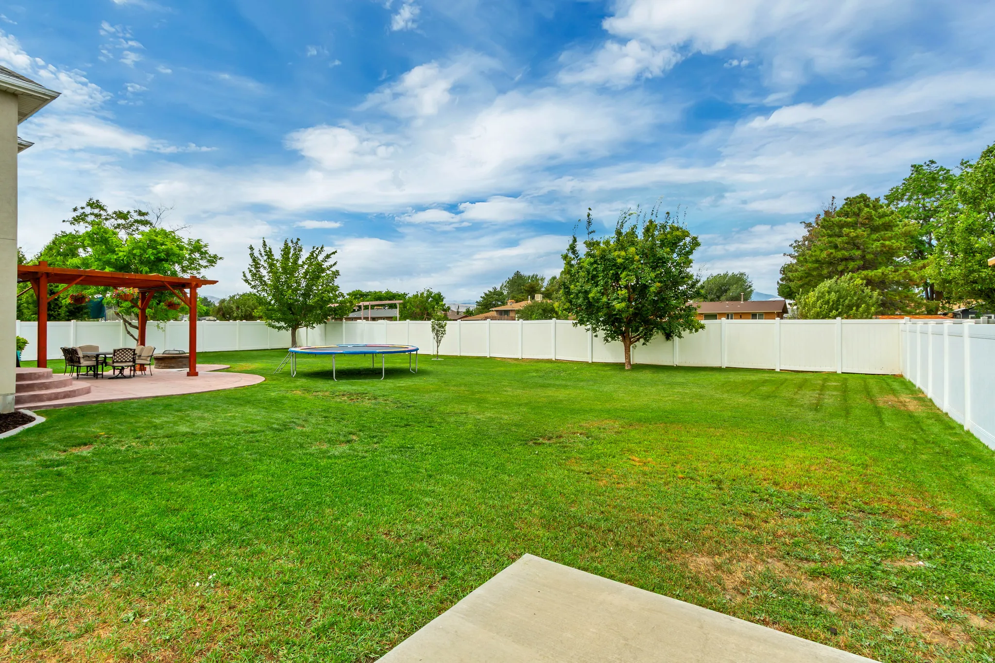 Expansive rear yard featuring a large green lawn, a white privacy fence, mature trees, and a concrete patio with a wooden pergola