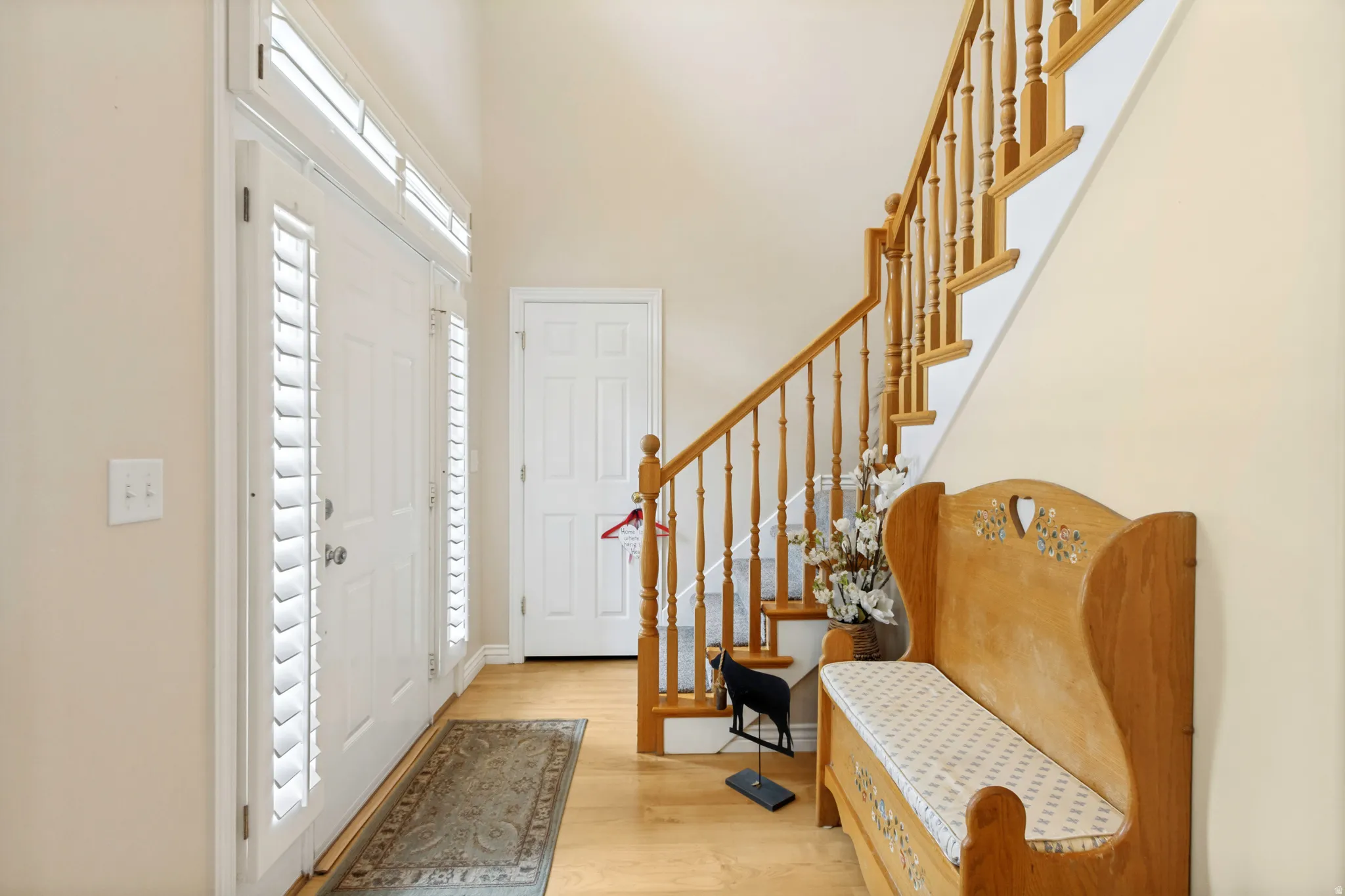 Entryway featuring a solid wood staircase with balusters, wood-finish flooring, and a front entry door with side lites and transom windows, all equipped with plantation shutters