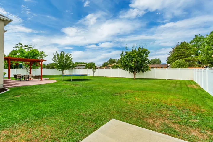 Expansive rear yard featuring a large green lawn, a white privacy fence, mature trees, and a concrete patio with a wooden pergola