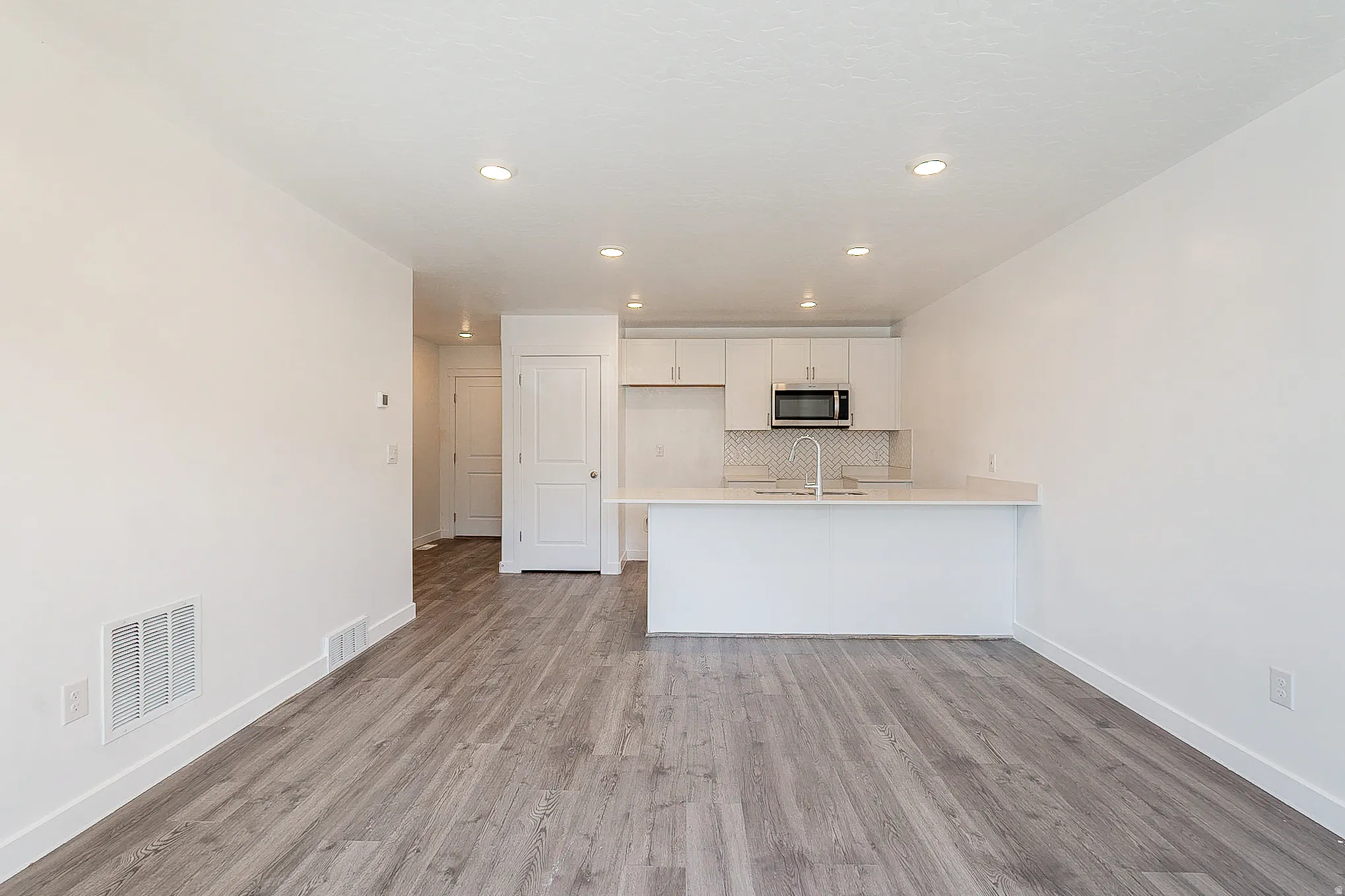 Open-concept living and kitchen area featuring wood-finish flooring throughout, recessed ceiling lighting, and a white cabinetry kitchen with stainless steel appliances and a breakfast bar