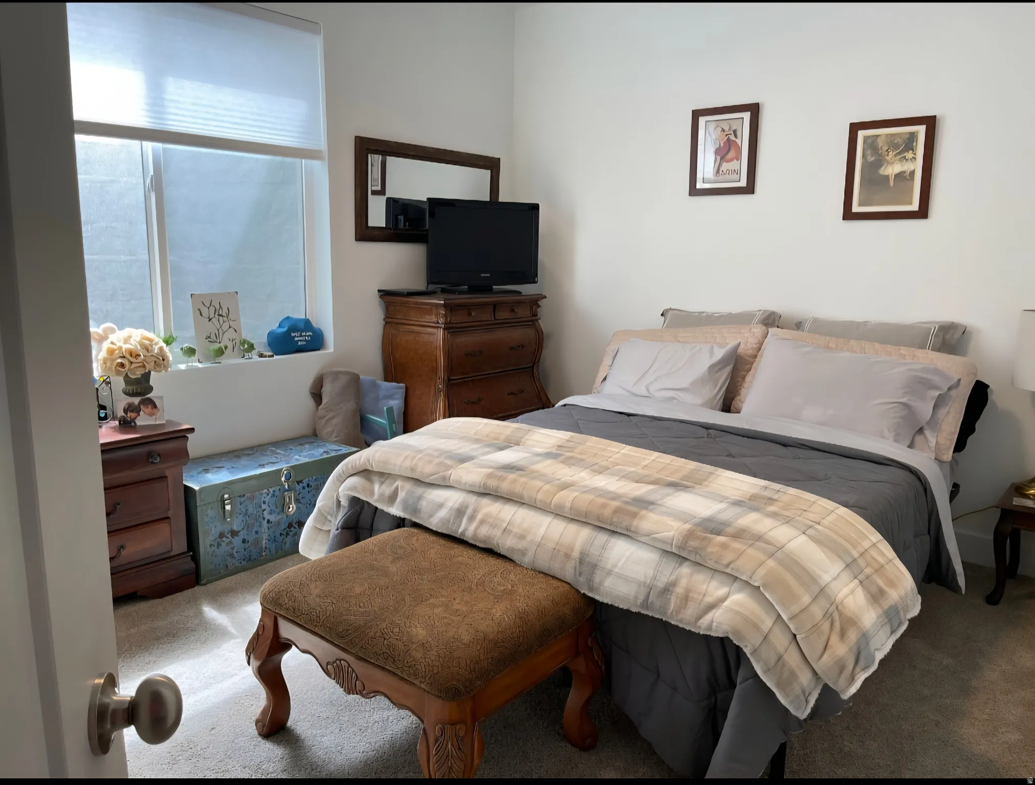 Bright bedroom featuring a large window with natural light, wood-finish dresser, framed wall art, plush area rug, and generous bed space with neutral upholstered headboard and coordinating bedding