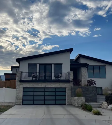 Modern home  featuring a striking contemporary facade with mixed stone and stucco exterior, dark-framed multi-pane glass doors and windows, a steel-panel garage door with horizontal detailing, a front deck with metal railings, and clean-lined architectura