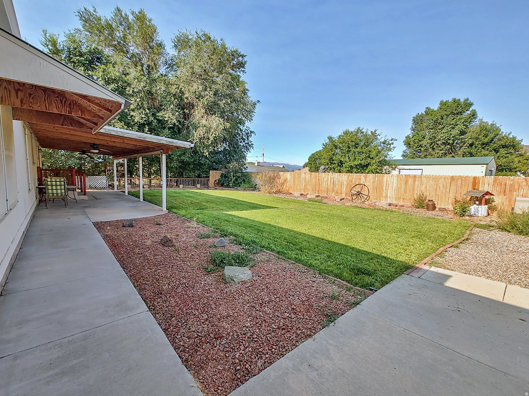 Expansive backyard featuring a large concrete patio, covered porch with exposed wood ceiling and ceiling fan, lush green lawn, mature trees, and a wooden privacy fence