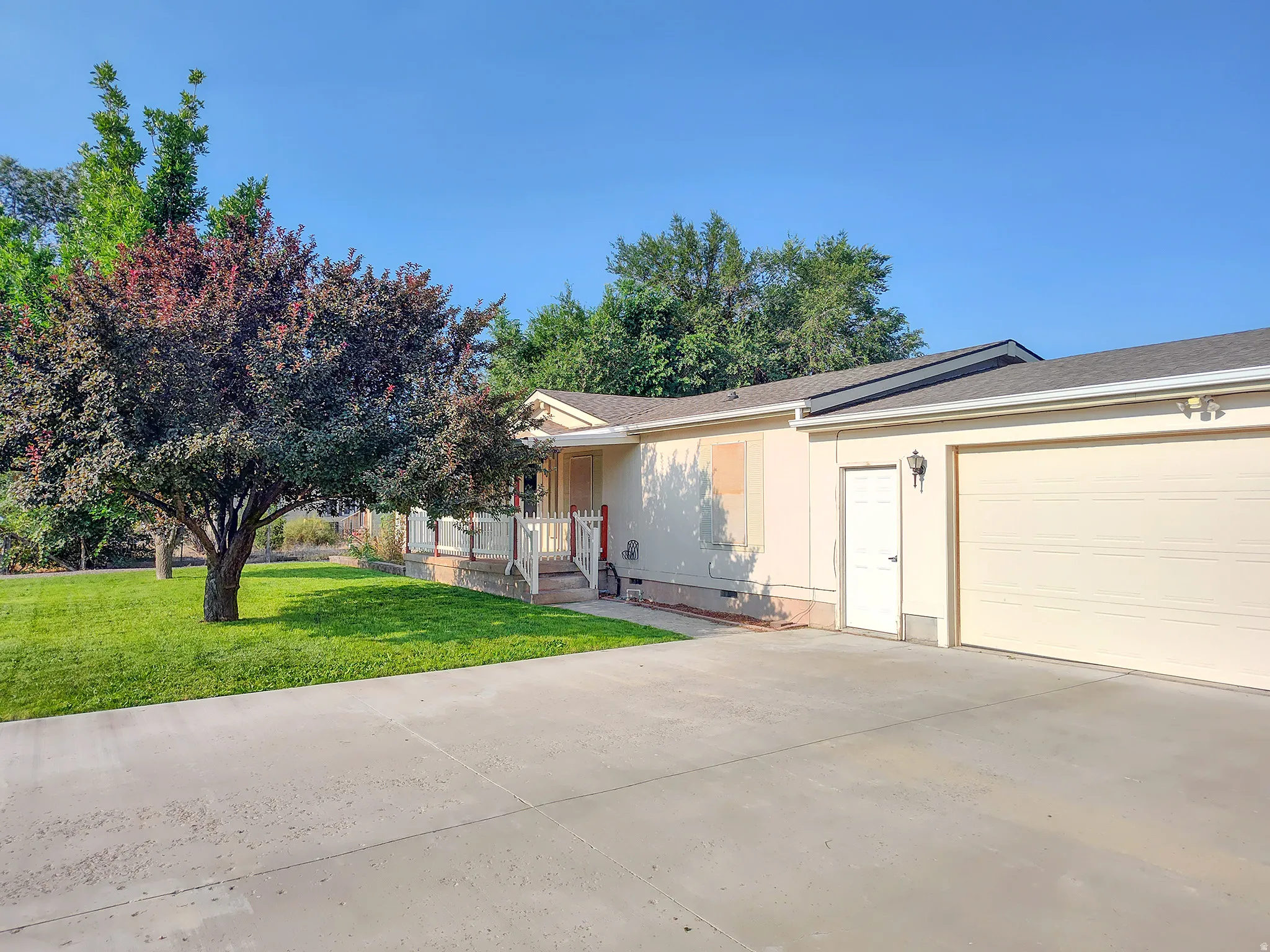 Charming single-story home featuring a spacious concrete driveway with dual-car garage access, mature landscaping with established trees providing natural shade, and a covered front porch entry