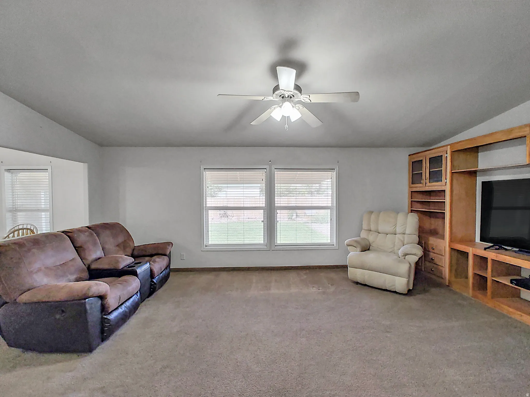Spacious living room featuring neutral carpeting, ceiling fan with integrated lighting, and large dual-pane windows with plantation shutters