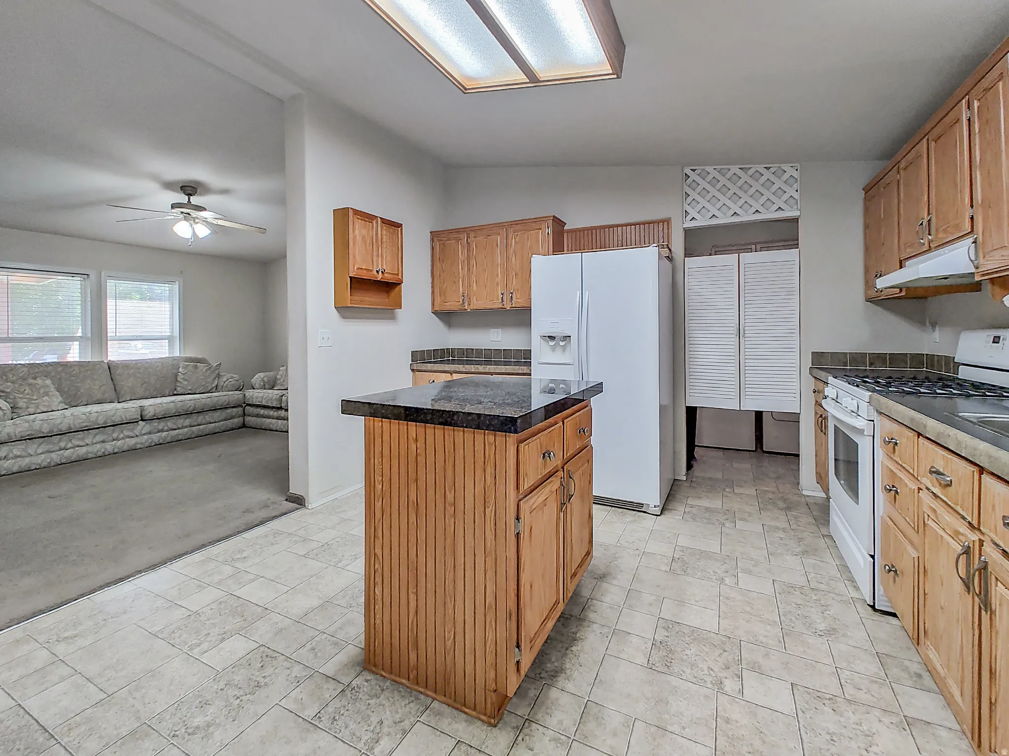 Spacious kitchen featuring wood-finish cabinetry, a central island with black countertop, white appliances, tile flooring, and abundant natural light from skylights