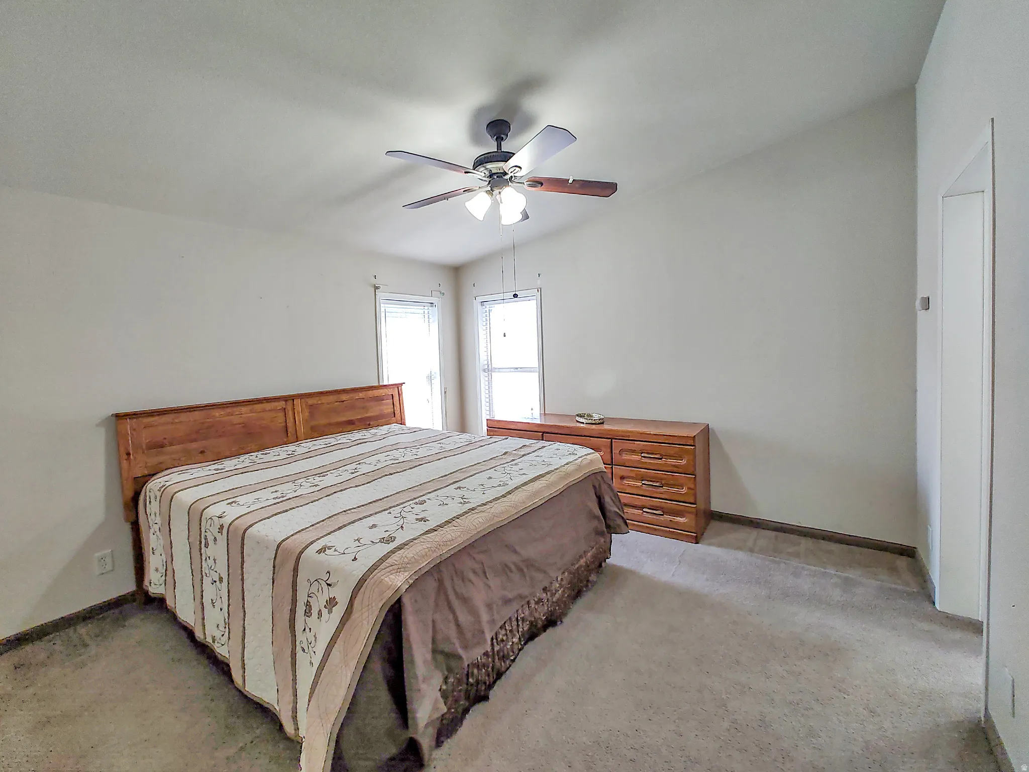 Spacious bedroom featuring a wood-finish headboard and nightstand, ceiling fan with integrated lighting, dual windows with natural light, and neutral carpeting throughout