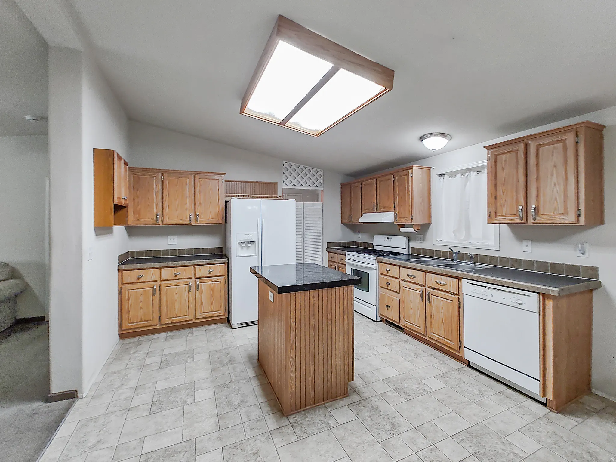 Well-appointed kitchen featuring wood-finish cabinetry, a central island with a dark countertop, white appliances, and a skylight for natural illumination