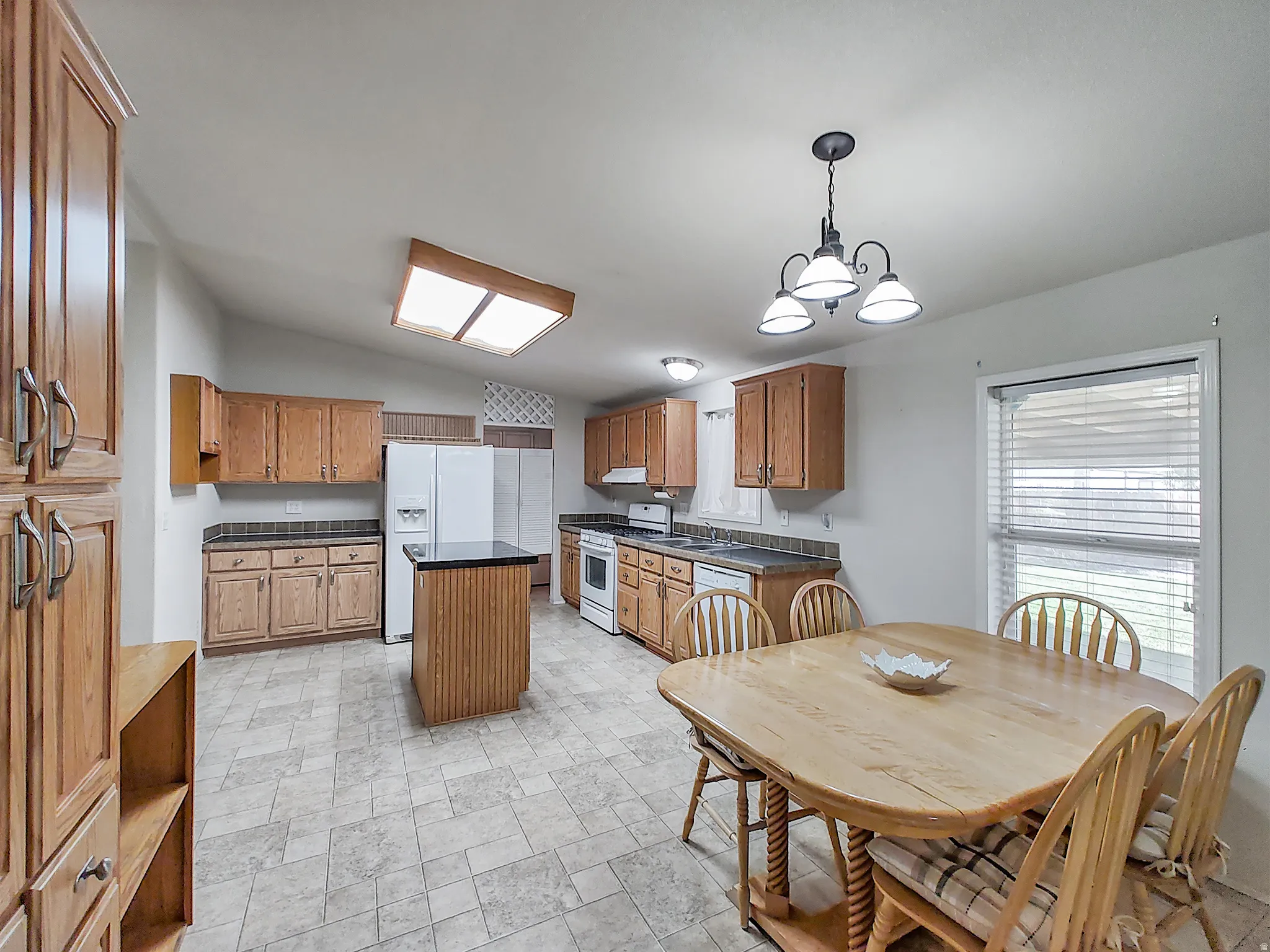 Spacious kitchen and dining area featuring wood cabinetry, white appliances, tile flooring, a central island with seating, pendant lighting, recessed ceiling fixtures, and a dining table with direct access to the adjoining room through glass doors with pl