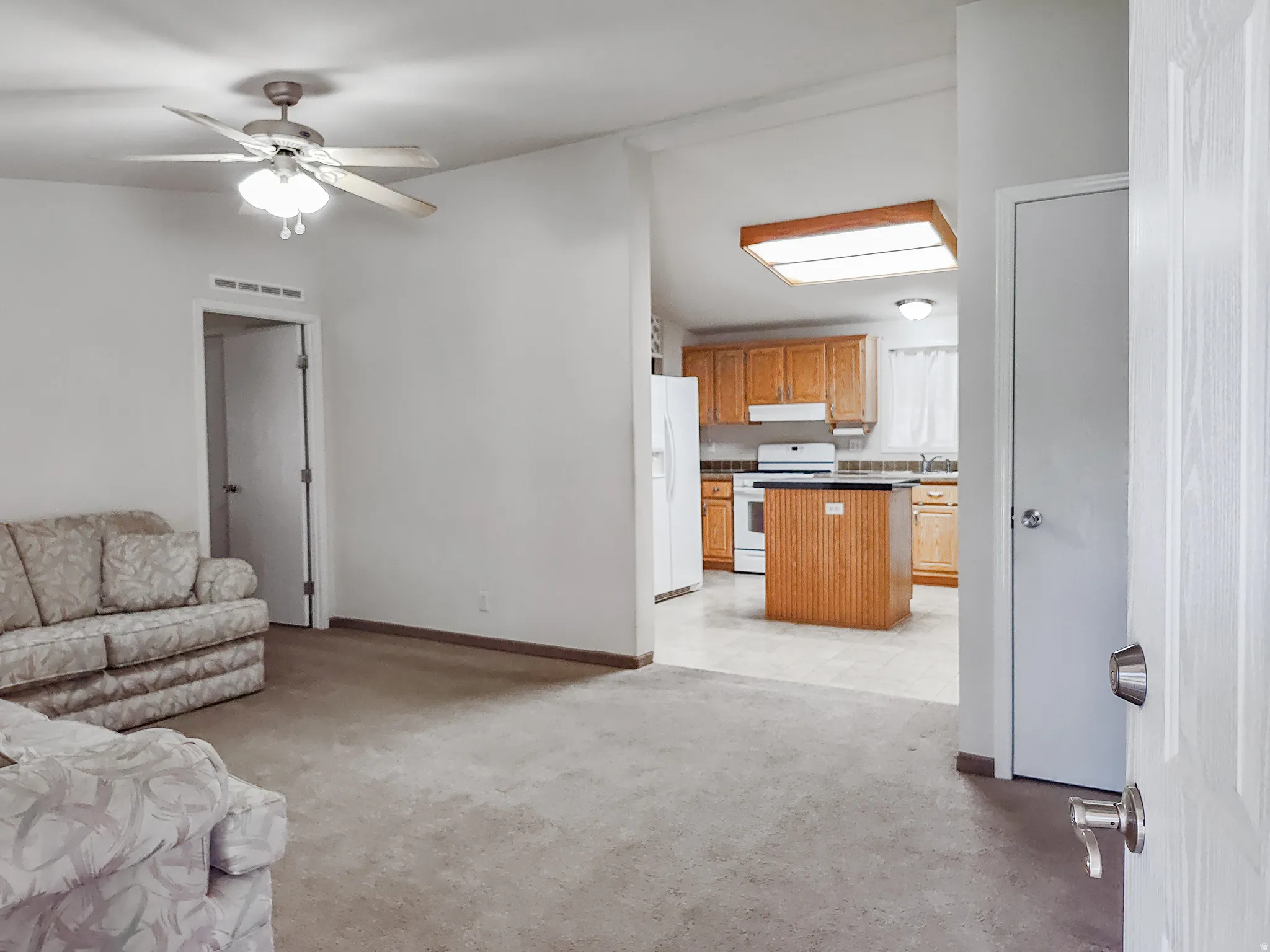 Living area with carpeting and a ceiling fan, offering a partial view of the kitchen