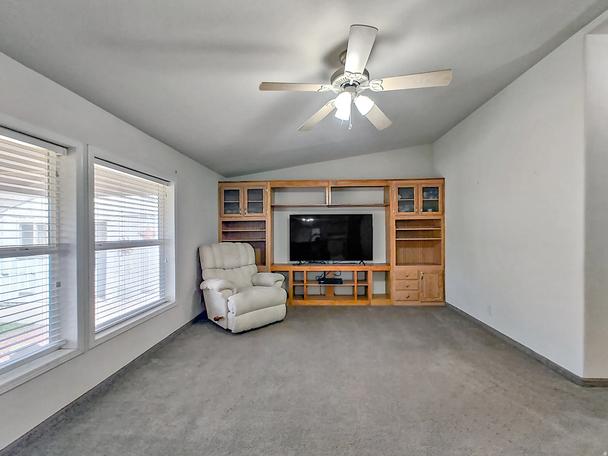 Spacious entertainment room featuring wall-mounted flat-screen television with custom wood cabinetry and built-in shelving, ceiling fan with integrated lighting, carpet flooring, and expansive windows with horizontal blinds allowing natural light througho