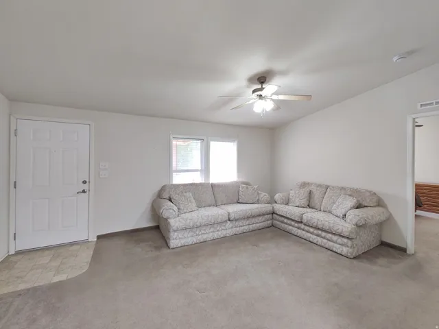 Living area featuring a ceiling fan with light fixture, neutral carpeting, and a white paneled entry door with hardware
