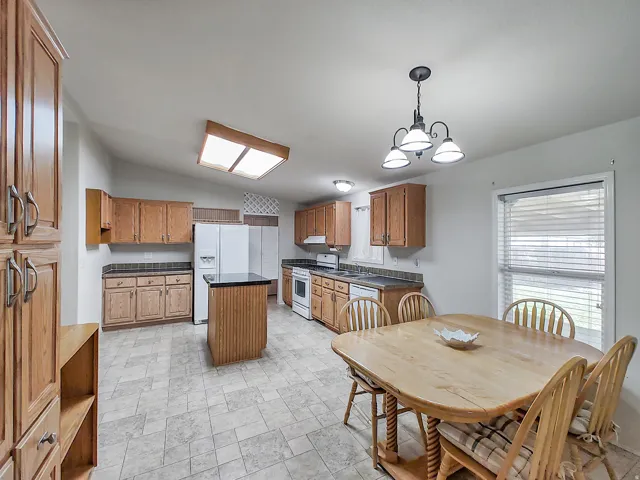 Spacious kitchen and dining area featuring wood cabinetry, white appliances, tile flooring, a central island with seating, pendant lighting, recessed ceiling fixtures, and a dining table with direct access to the adjoining room through glass doors with pl