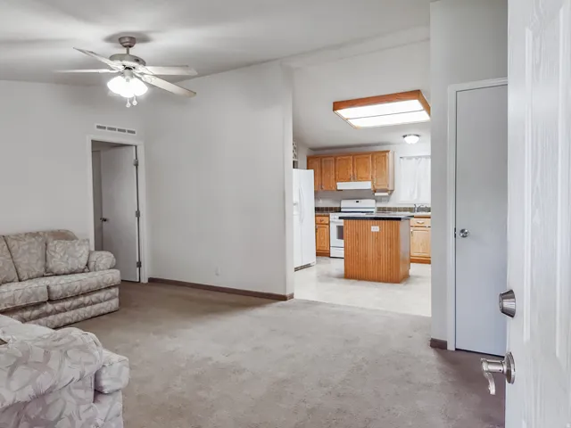 Living area with carpeting and a ceiling fan, offering a partial view of the kitchen