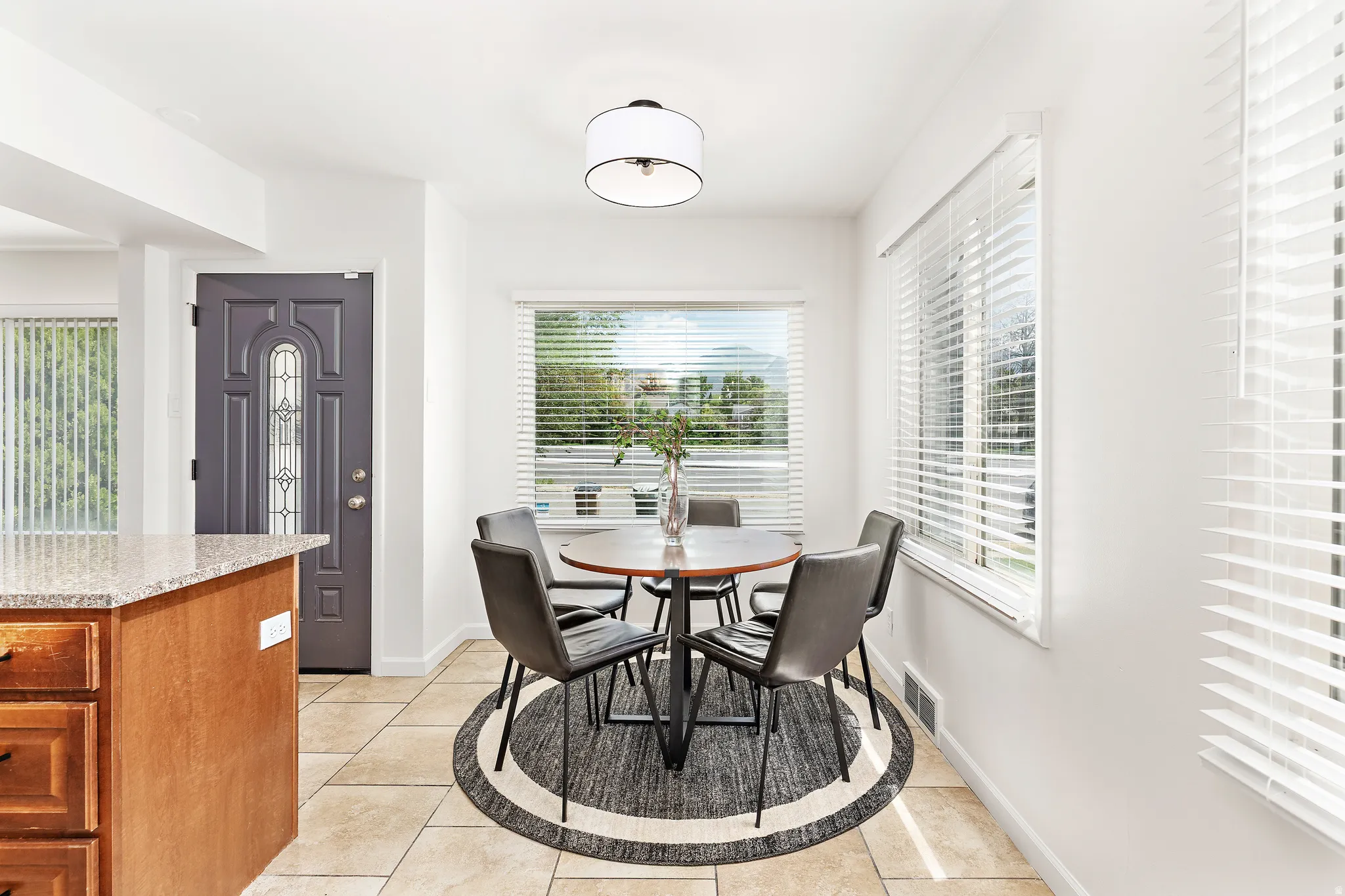 Bright dining area featuring tile flooring, multiple windows with blinds, a drum pendant light fixture, and a decorative front entry door with glass inserts