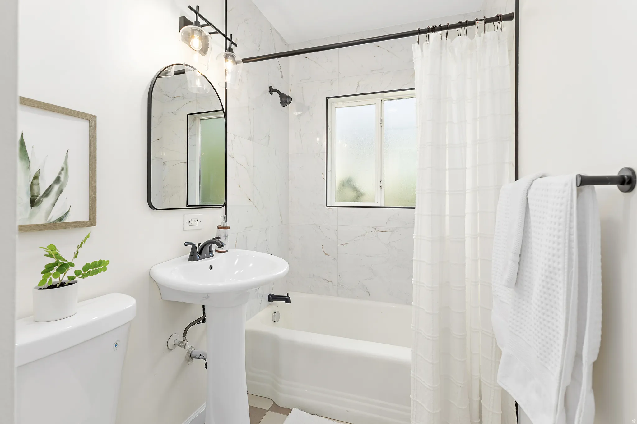 Bathroom featuring a pedestal sink, black-finish fixtures, an arched mirror, an integrated tub/shower with tile surround, and a frosted window