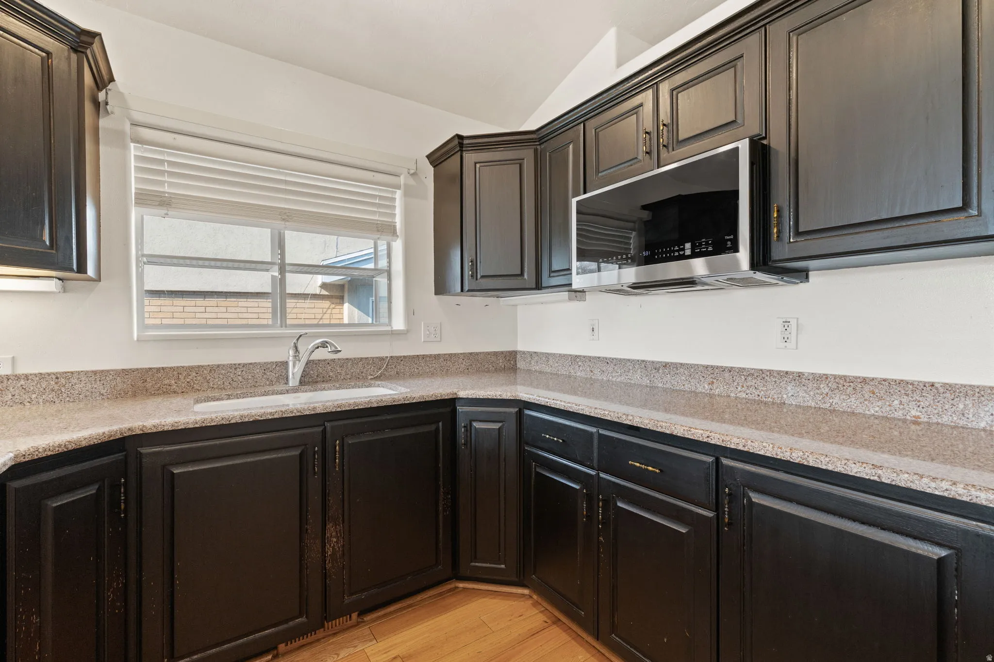 Dark-finish cabinetry with panel doors, integrated stainless steel microwave, light-toned countertops, and a single basin sink with a gooseneck faucet