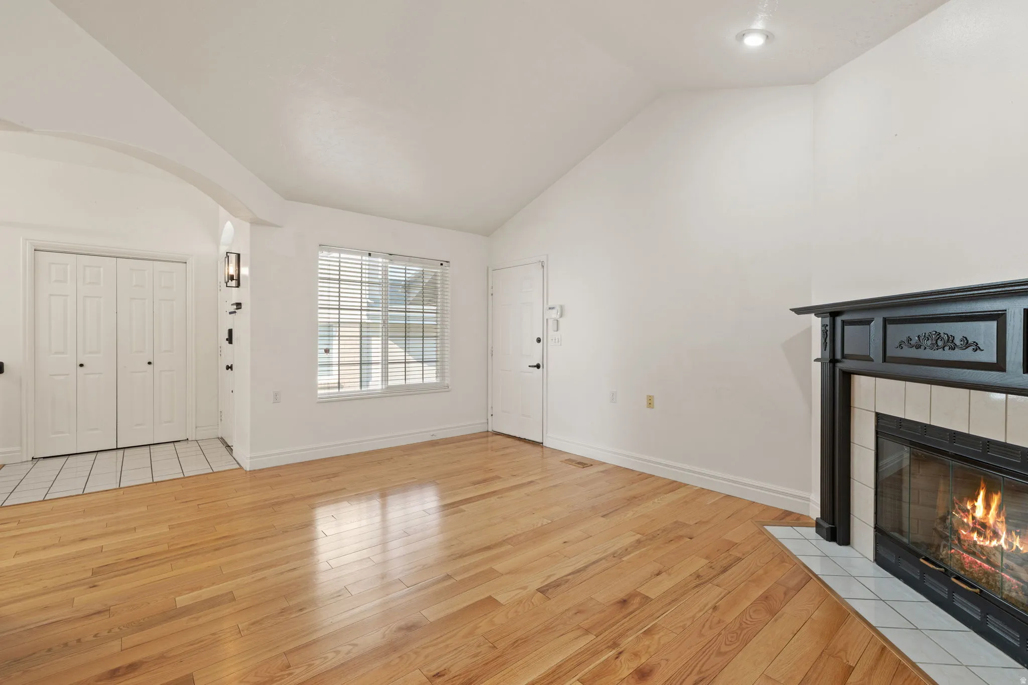Vaulted ceiling, wood-finish flooring, tiled entry, window with blinds, and a fireplace with a decorative mantel and tiled surround