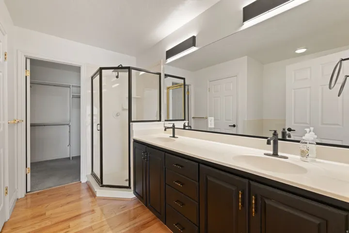 Dual vanity with integrated sinks and black base cabinetry, corner glass shower enclosure, wood-finish flooring, and a walk-in closet with carpet