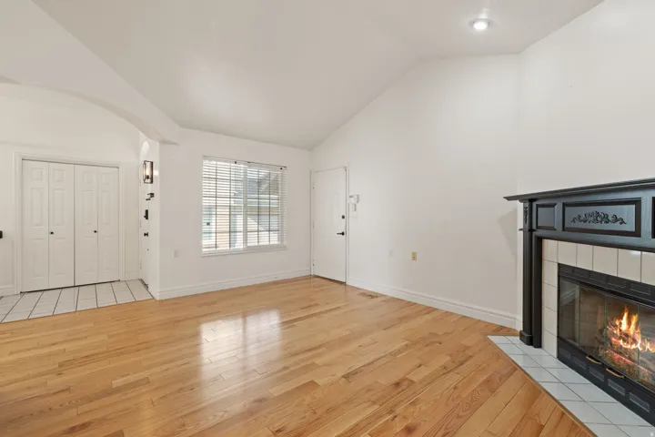 Vaulted ceiling, wood-finish flooring, tiled entry, window with blinds, and a fireplace with a decorative mantel and tiled surround