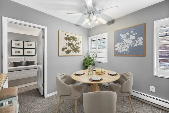 Ceiling fan with integrated lighting, two windows with plantation shutters, baseboard heating, and an open doorway leading to an additional room