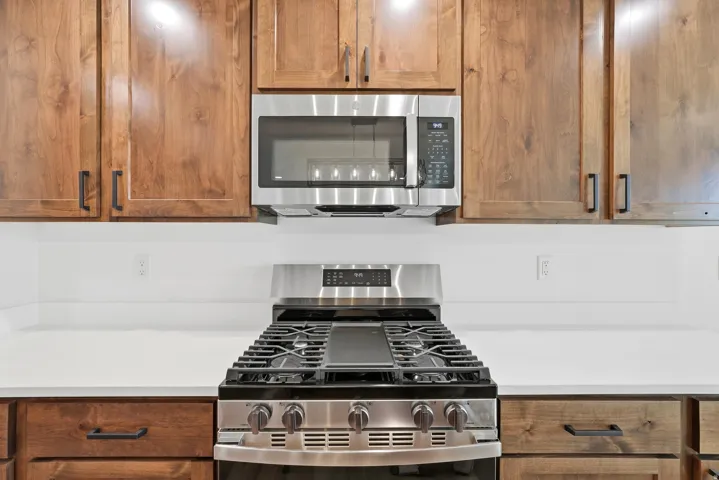 Stainless steel gas range with griddle, stainless steel over-the-range microwave, wood-finish cabinetry with matte black hardware, and solid surface countertops