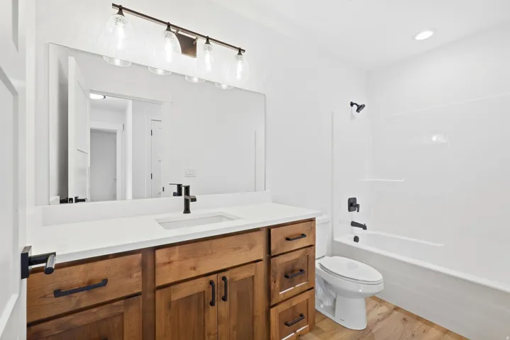 Wood-finish flooring, wood vanity with dark hardware, white countertop with integrated sink, dark faucet, and a full bathtub with dark fixtures